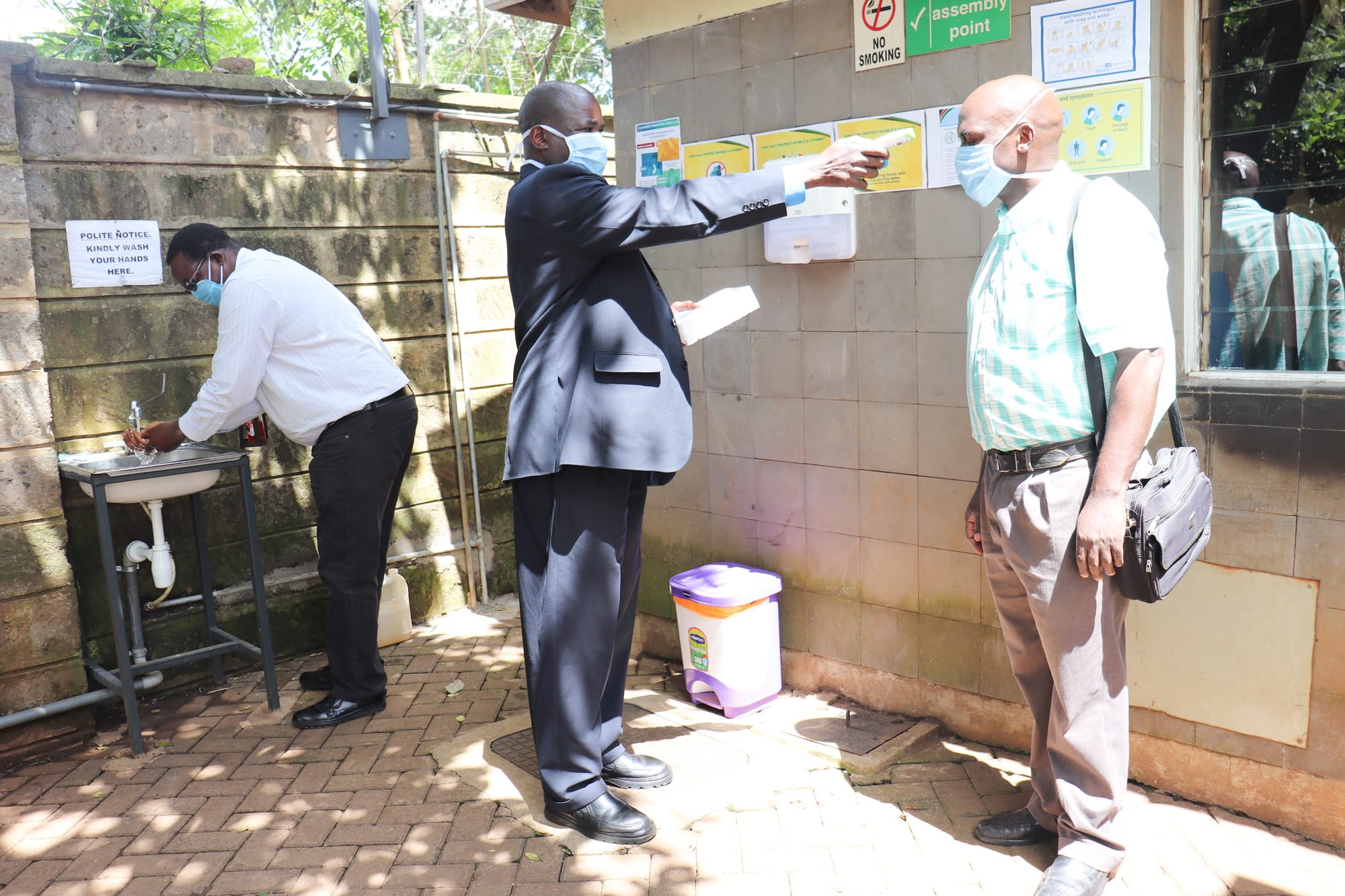 A man takes a temperature reading from a visitor at the Christian Health Association of Kenya gate, located in Nairobi, as a staff member washes his hands after having his temperature taken.