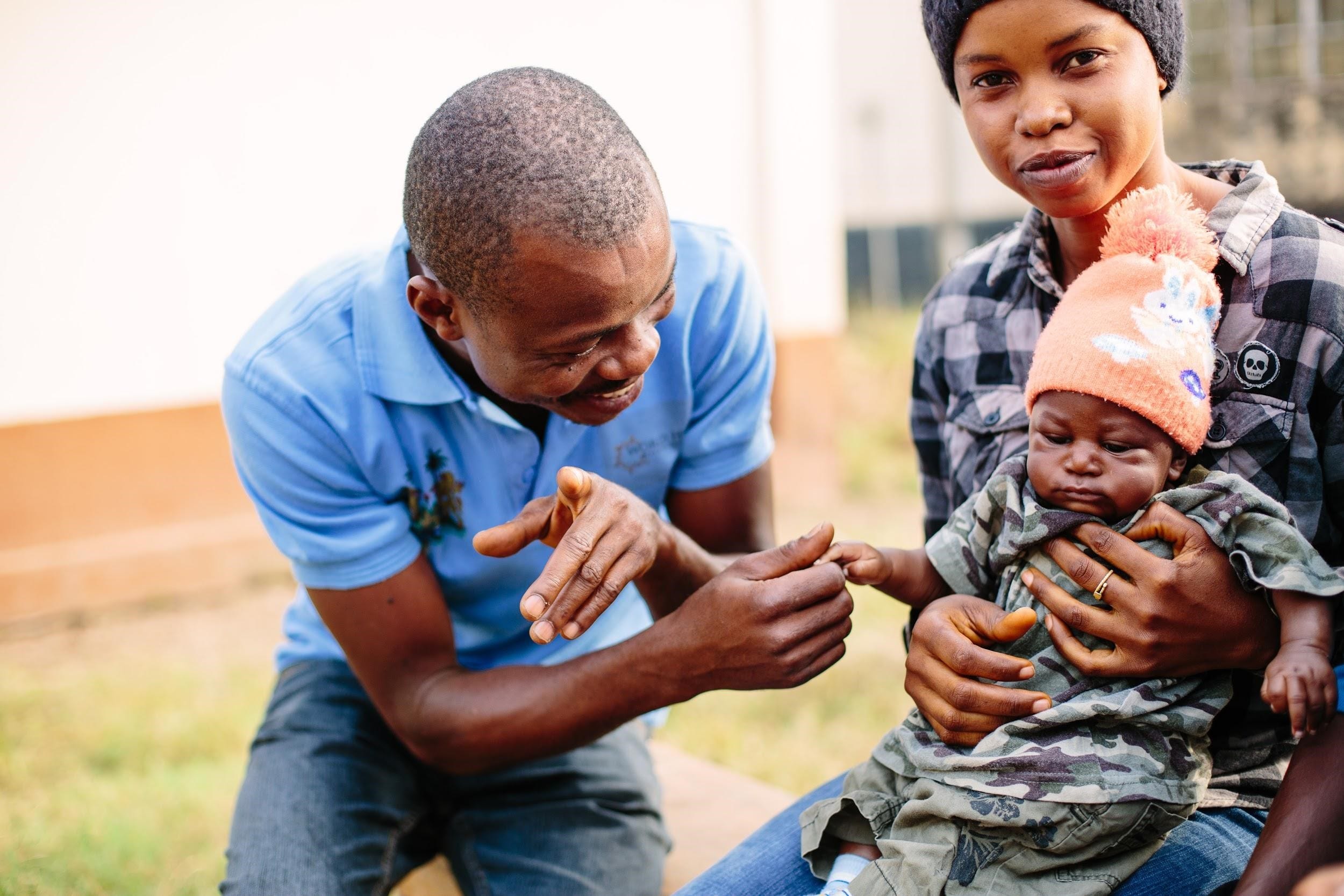 A male health worker smiles at an infant being held by his mother.