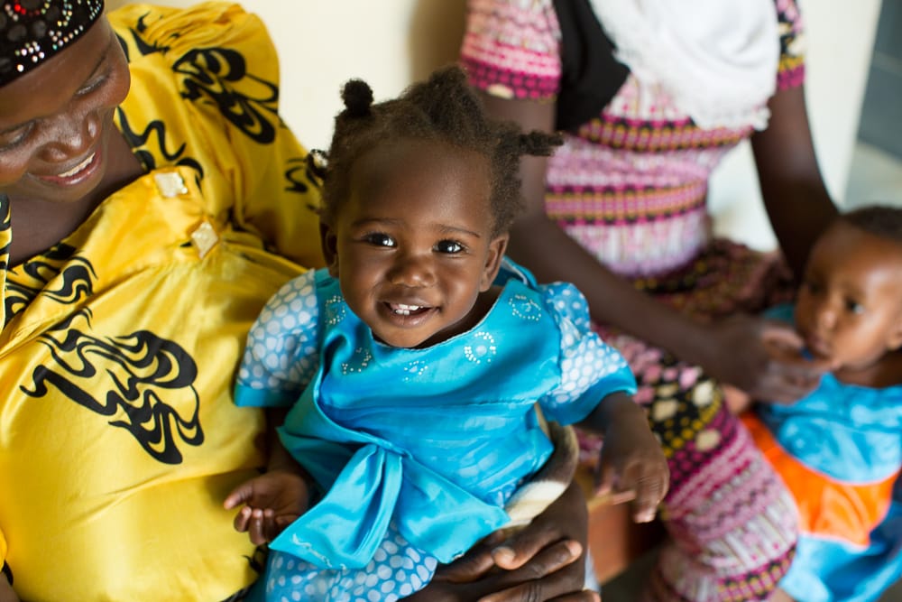 A young girl in Uganda in a blue dress sits on her mother's lap.