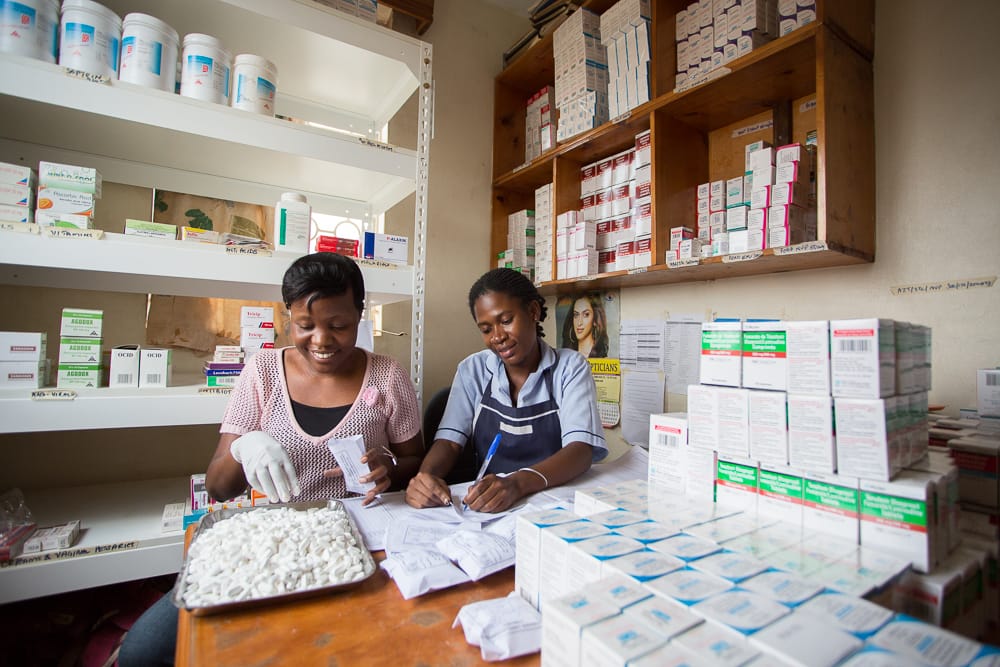 Two women sit at a table working in a pharmacy in a health facility in Uganda.
