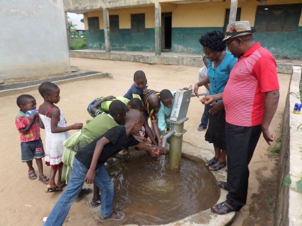 A woman and man stand near a water pump surrounded by children